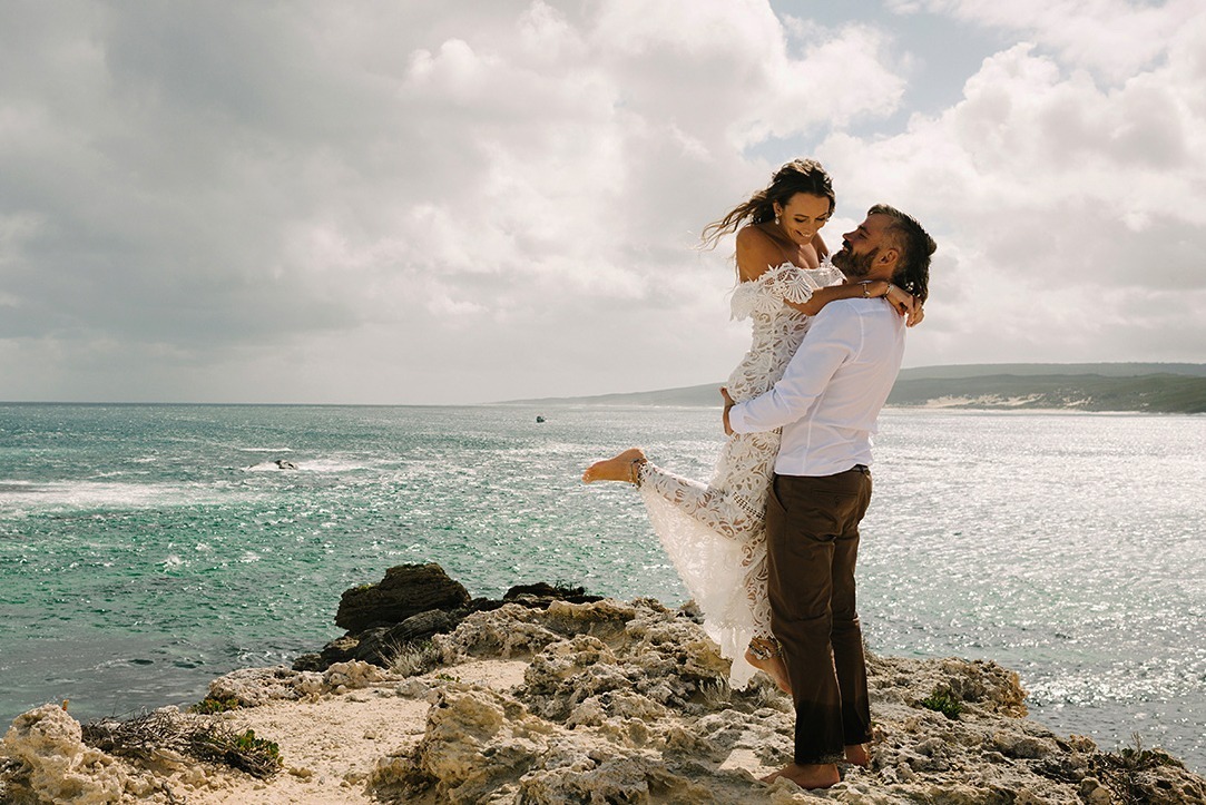 Groom lifting his bride off the ground as they kiss on the granite cliff edge above Kilcarnup Beach, Margaret River, her white gown catching the wind with turquoise ocean, white sand and coastal dunes stretching out behind them