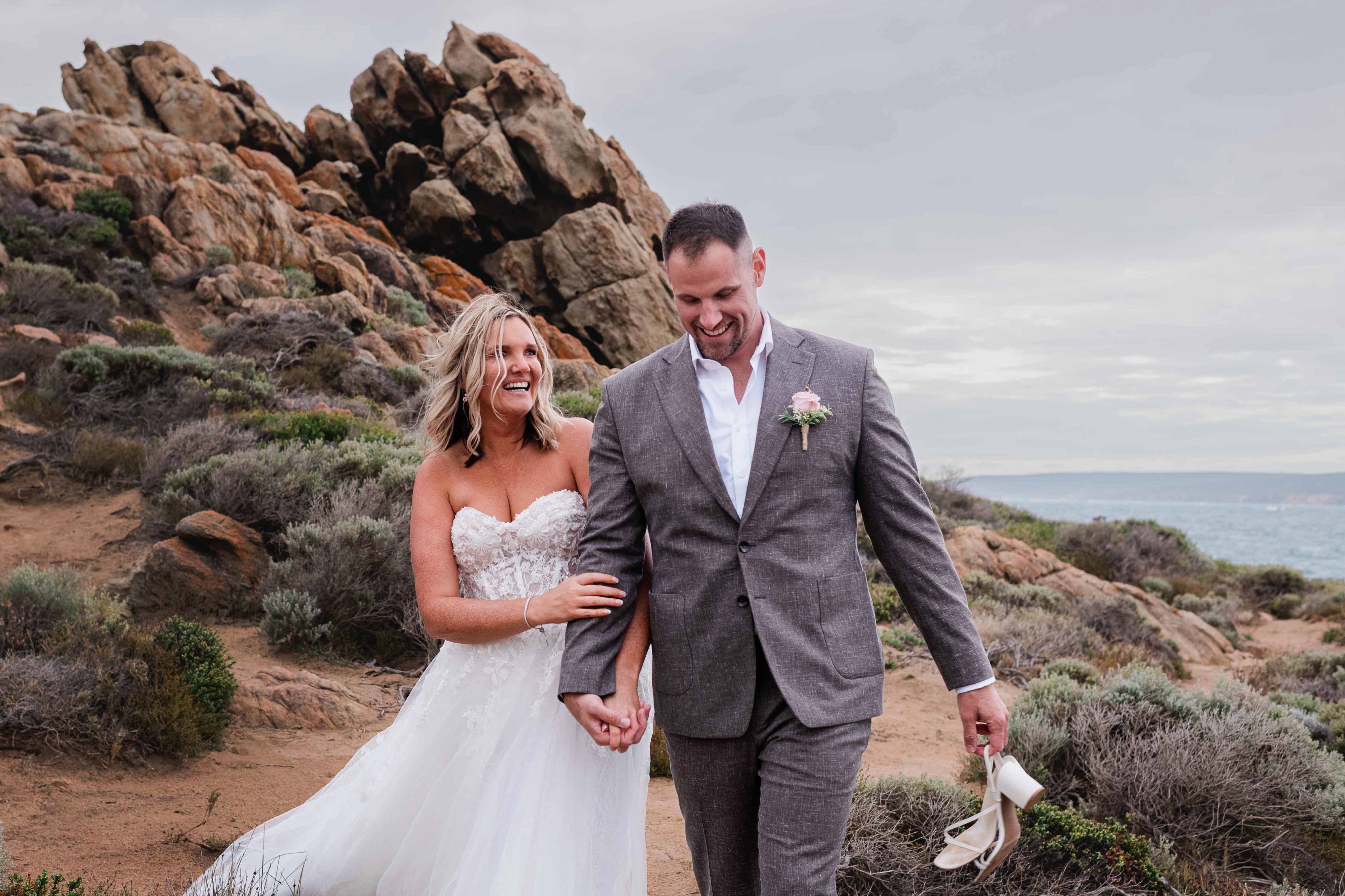 Bride in a strapless lace gown laughing with her groom in a grey linen suit as they walk hand in hand along the sandy coastal path at Canal Rocks, Yallingup, Margaret River region, with weathered granite outcrops and silver coastal heath rising behind them and the Indian Ocean off to the right