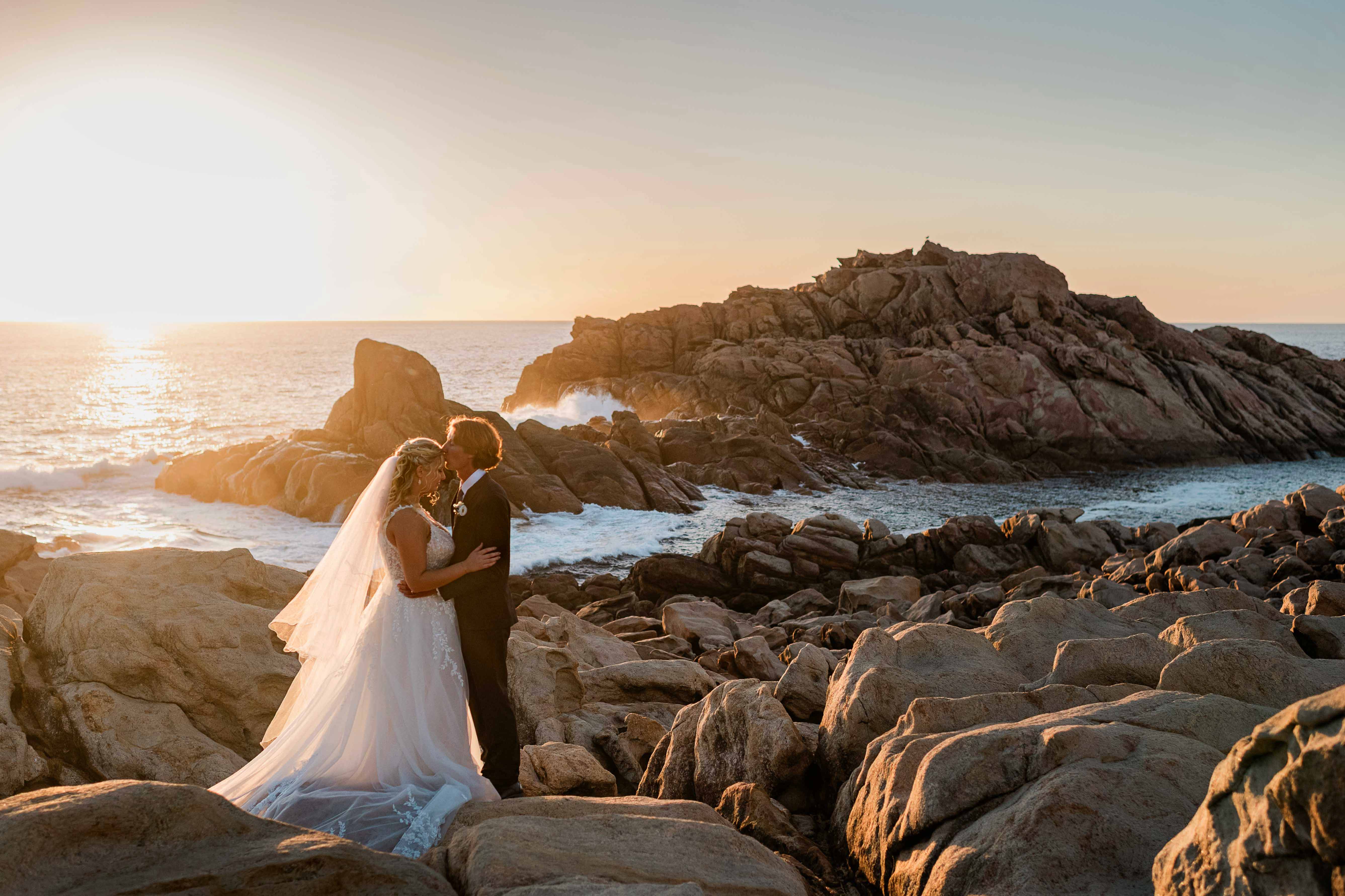 Groom kissing his bride on the forehead at sunset on the granite boulders at Canal Rocks, Yallingup, in the Margaret River region, veil trailing across the rocks as waves crash into the headland and golden light spreads across the Indian Ocean behind them