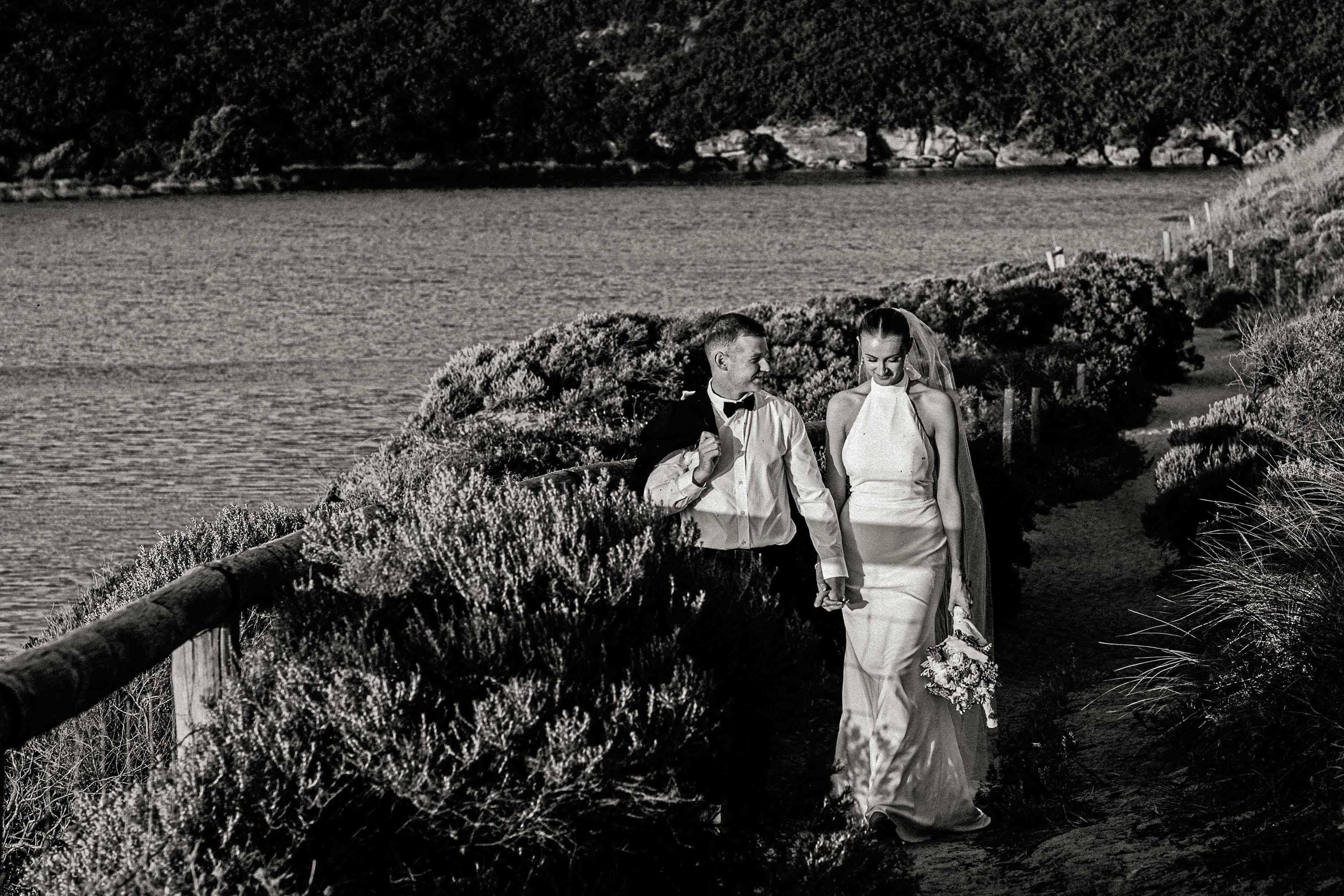 Black and white portrait of a bride in a high-neck silk gown and long veil walking hand in hand with her groom along the timber-railed coastal path above the river at Margaret River Mouth Beach, coastal heath spilling over the fence and the still river water stretching behind them