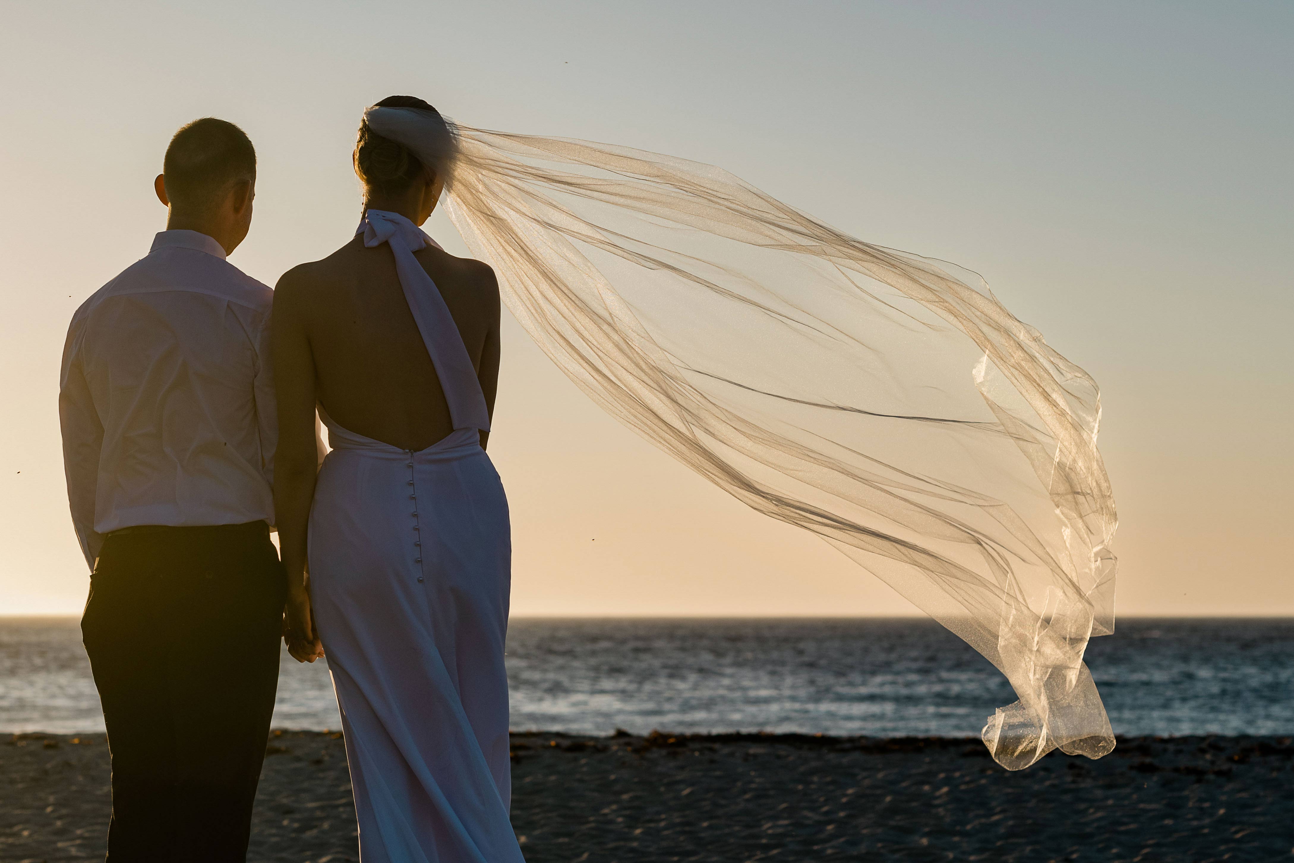 Bride in a backless halter-neck silk gown standing hand in hand with her groom at the shoreline of Margaret River Mouth Beach at sunset, her long tulle veil catching the wind and streaming out horizontally across the golden sky above the Indian Ocean