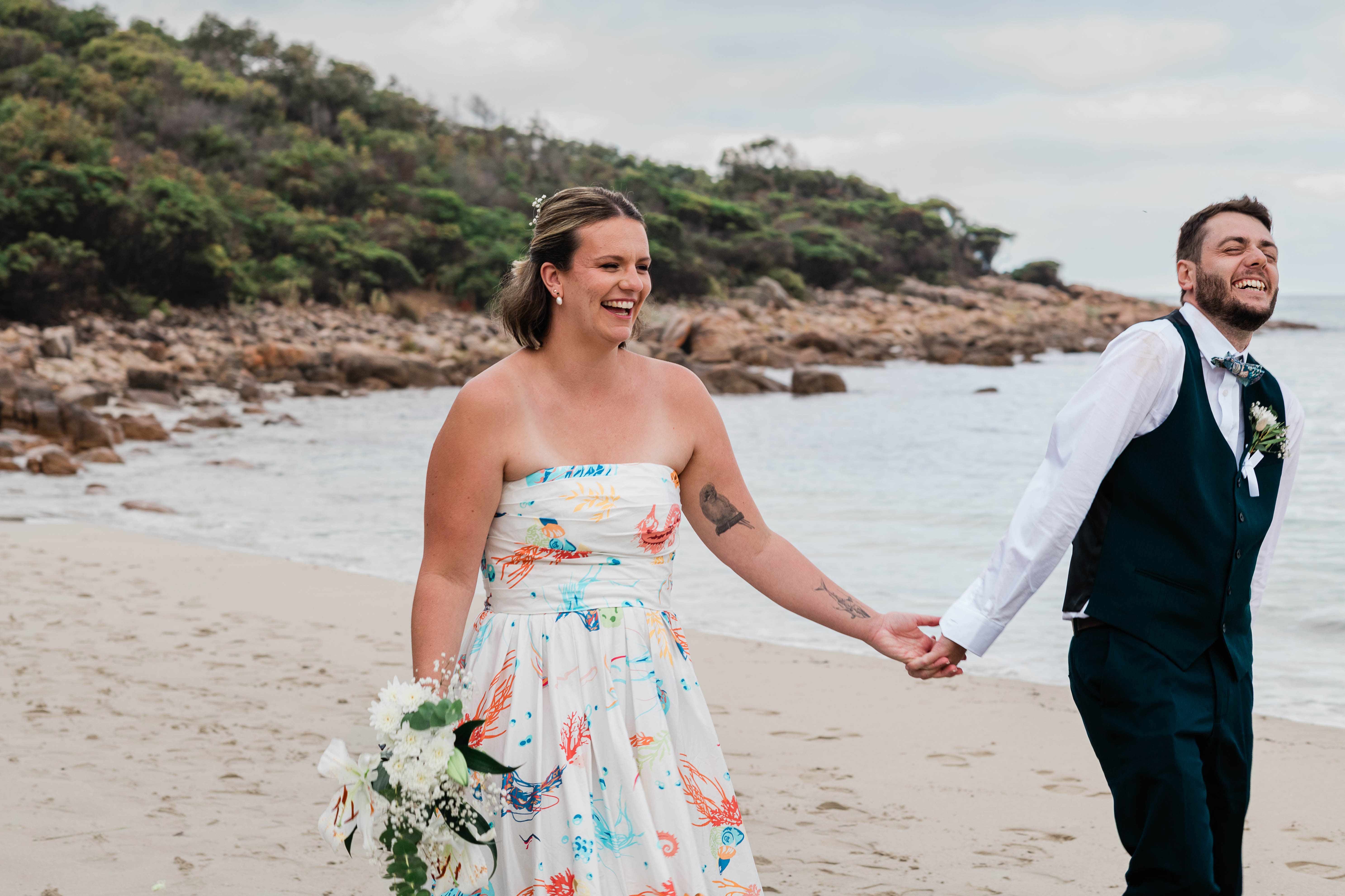 Bride in a hand-painted reef-print gown holding hands with her groom in a navy waistcoat and blue bow tie, both laughing as they walk along the sand at Meelup Beach, Margaret River, with granite headland and turquoise water behind them