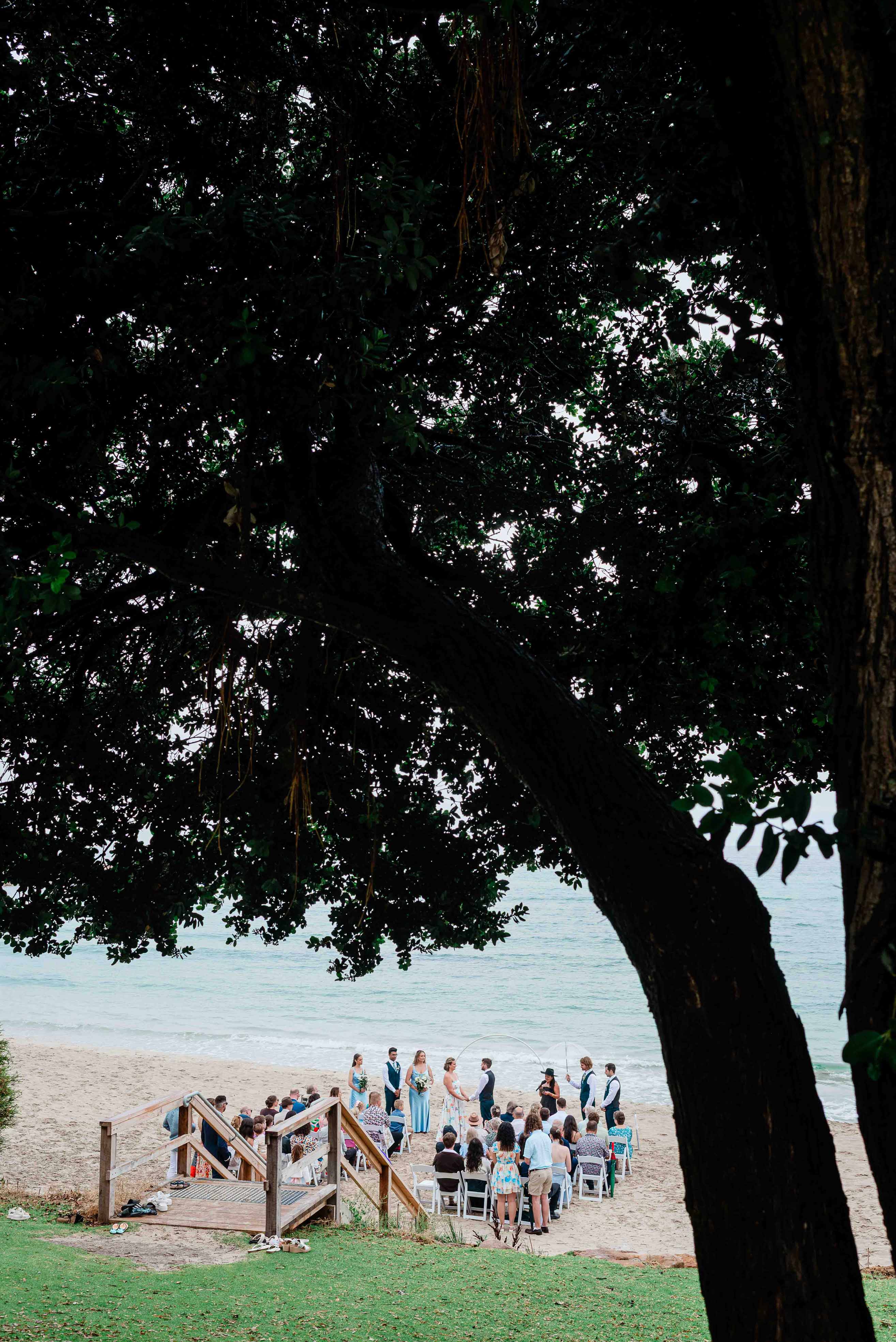 Wedding ceremony gathered on the sand at Meelup Beach, Margaret River, framed through the sprawling dark canopy of an old peppermint tree, with guests seated on white chairs, the wedding party at a clear arbour and the calm waters of Geographe Bay behind