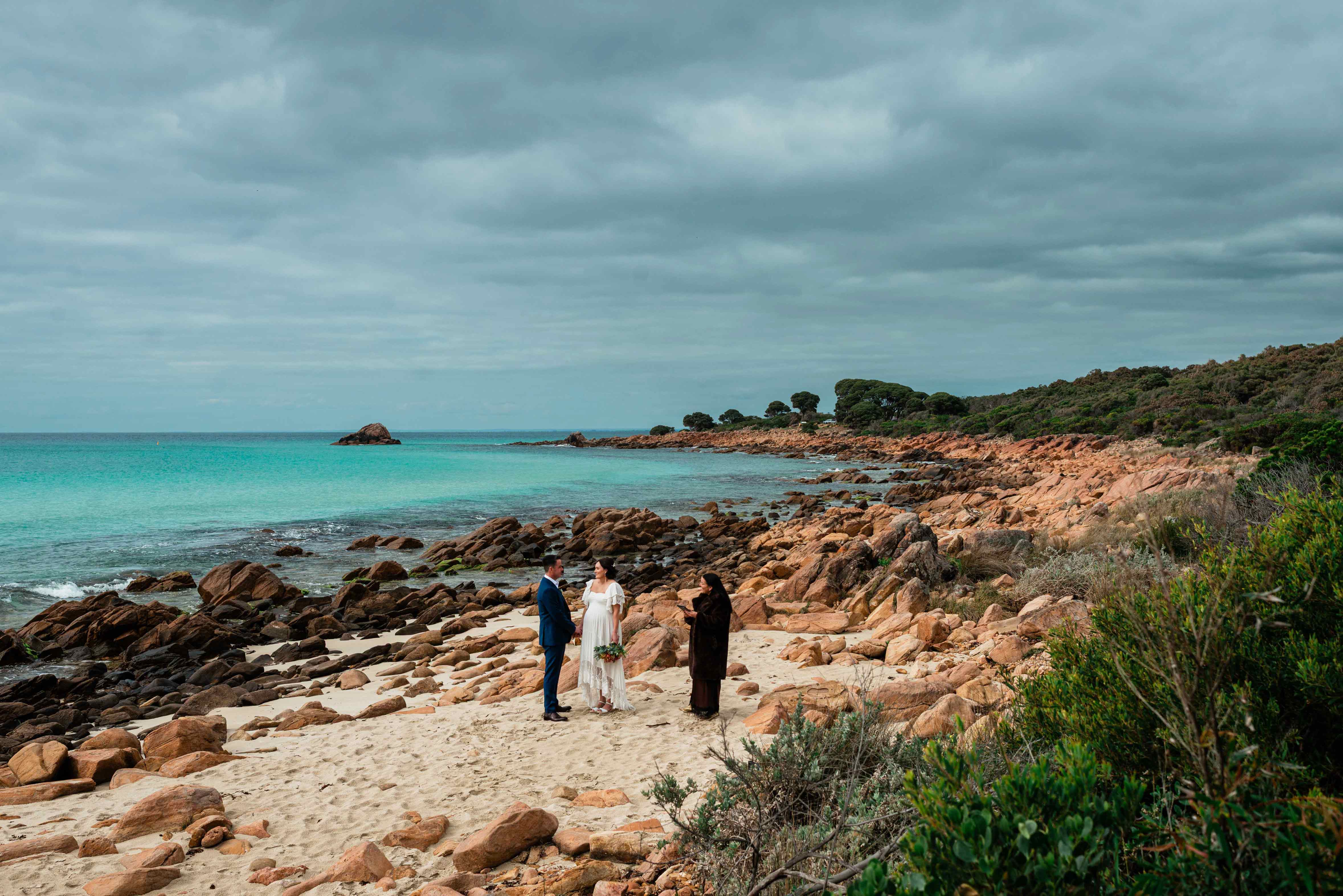 Bride and groom holding hands during their elopement ceremony with a celebrant on the white sand between orange granite boulders at Point Piquet, Margaret River, with the turquoise waters of Geographe Bay curving into the distance under a moody grey sky