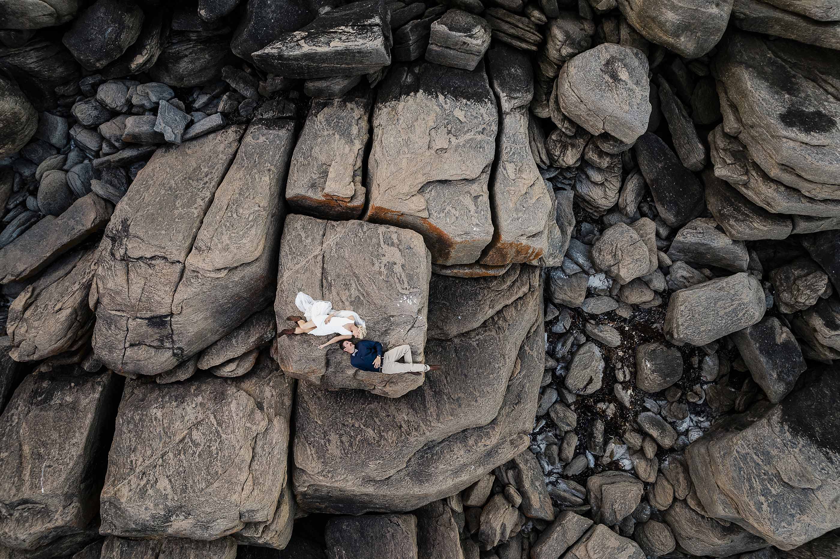 Aerial drone view of a bride and groom lying together on a huge weathered granite boulder at Shelley Cove, Margaret River, dwarfed by the cracked stacked rock formations of the coastline