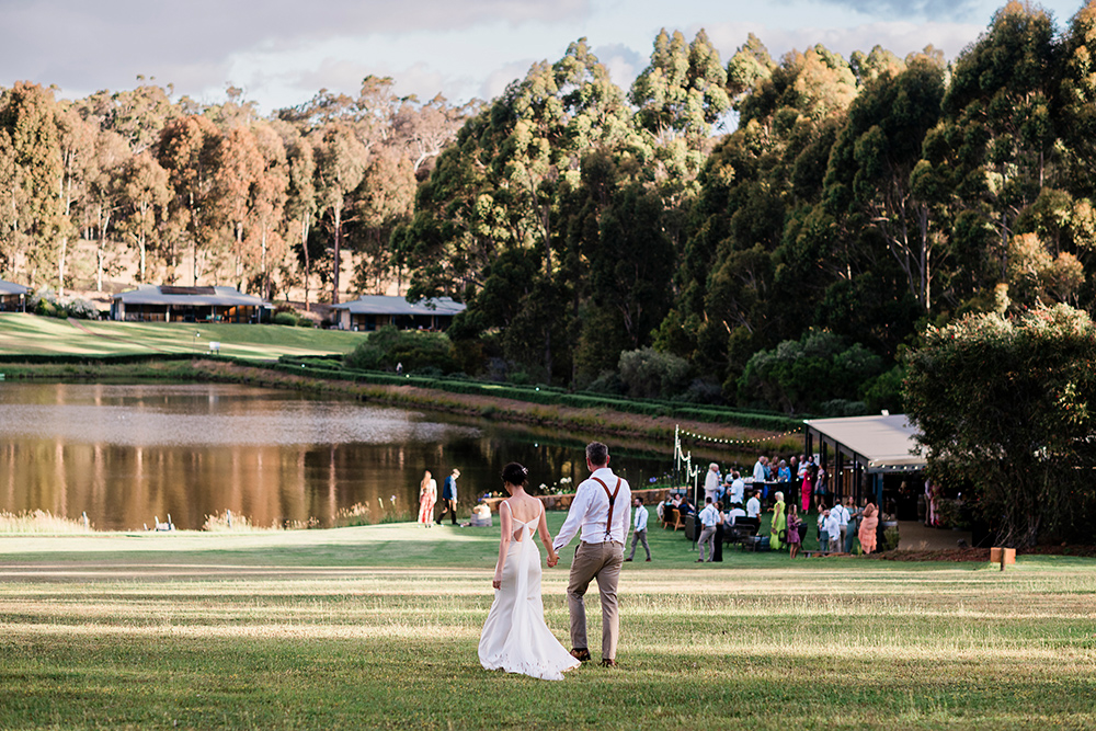 Bride and groom walking hand in hand across the lawn towards their lakeside reception at Tanah Marah, Margaret River, with guests gathered under festoon lights and karri forest reflecting in the dam at golden hour