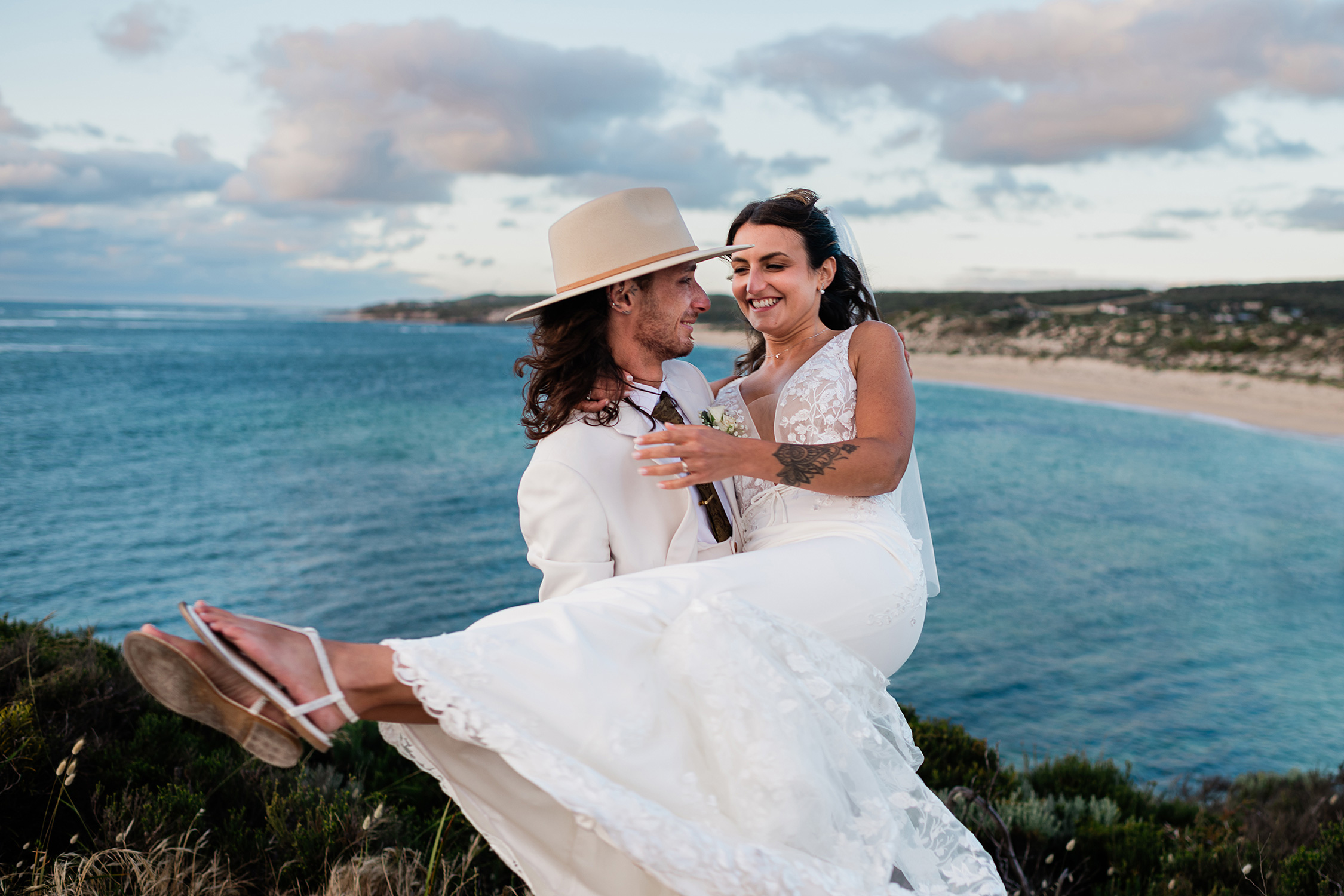 Groom in a wide-brim hat carrying the bride over the coastal cliff backdrop of Gnarabup Beach, Margaret River