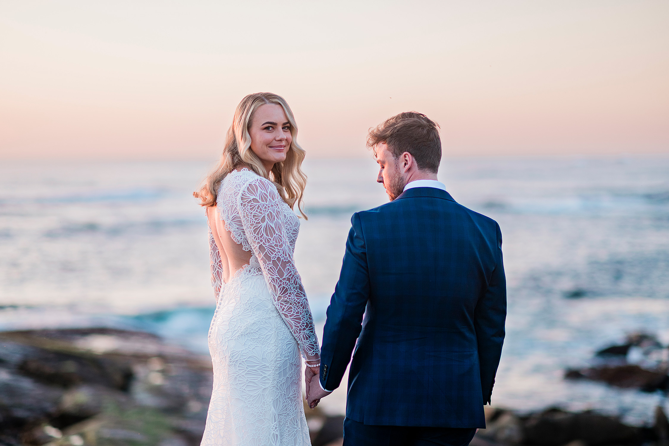Romantic Redgate Beach sunset elopement photo with bride looking back at camera and groom gazing at her, capturing a dramatic beach moment