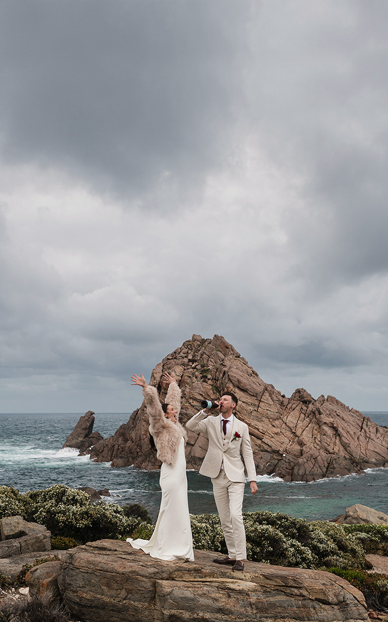 Eloping groom drinks champagne as bride lifts arms celebrating and laughing, with the dramatic Sugar Loaf Rock as their backdrop