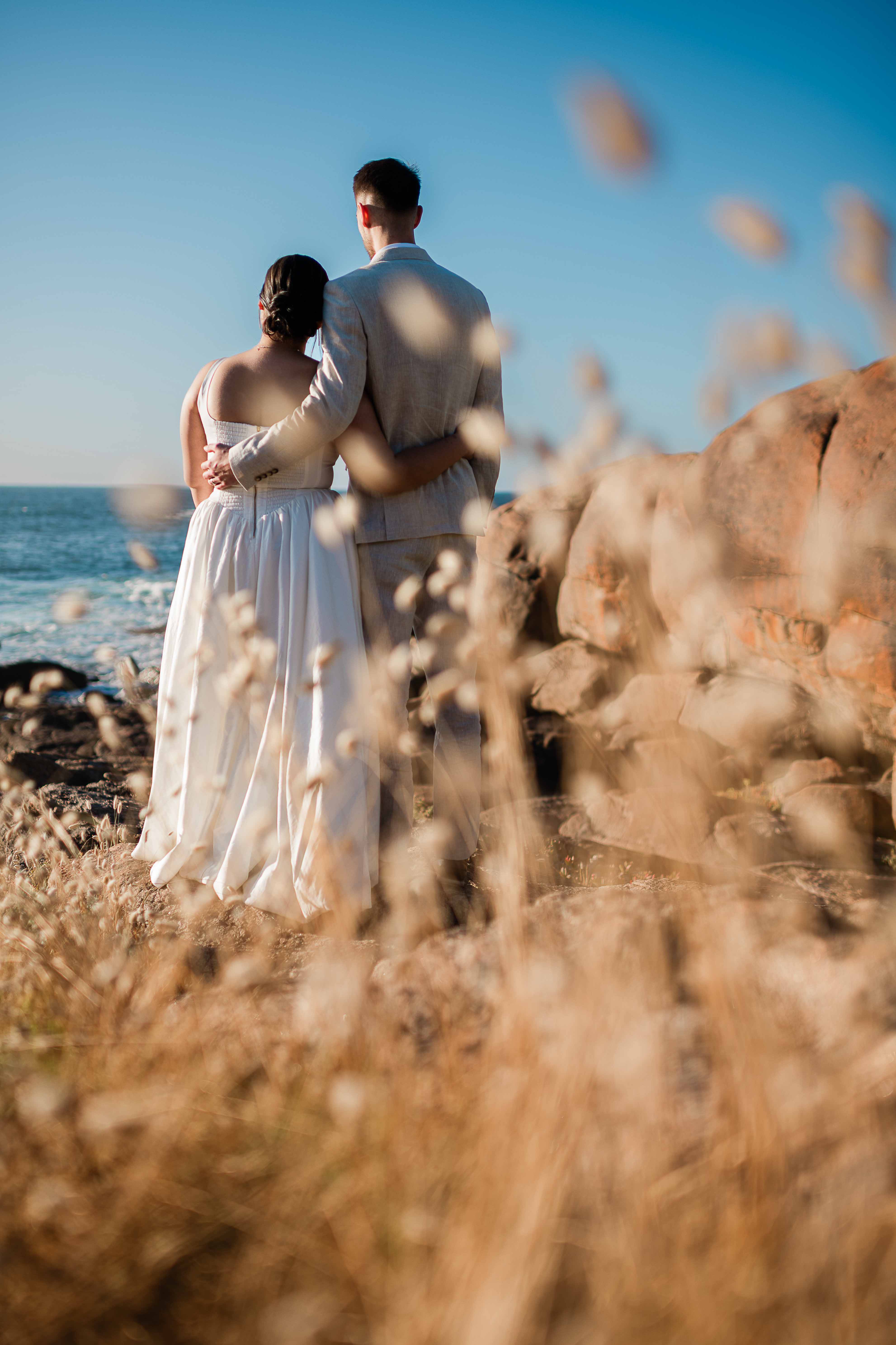 bride and groom framed by long golden grass look out to the ocean at golden hour