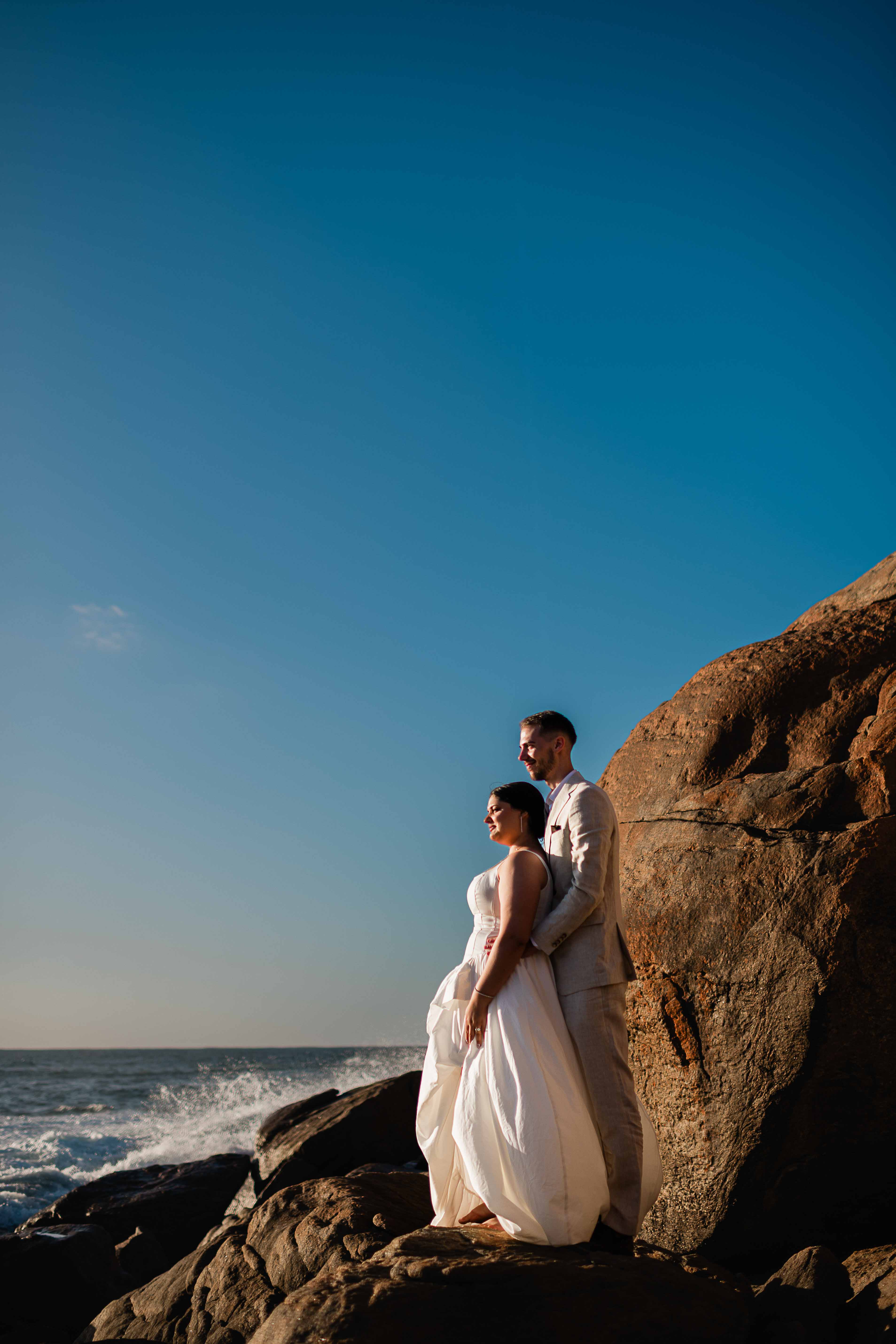 stunning light falls on bride and groom and large rocks with gorgeous ocean backdrop