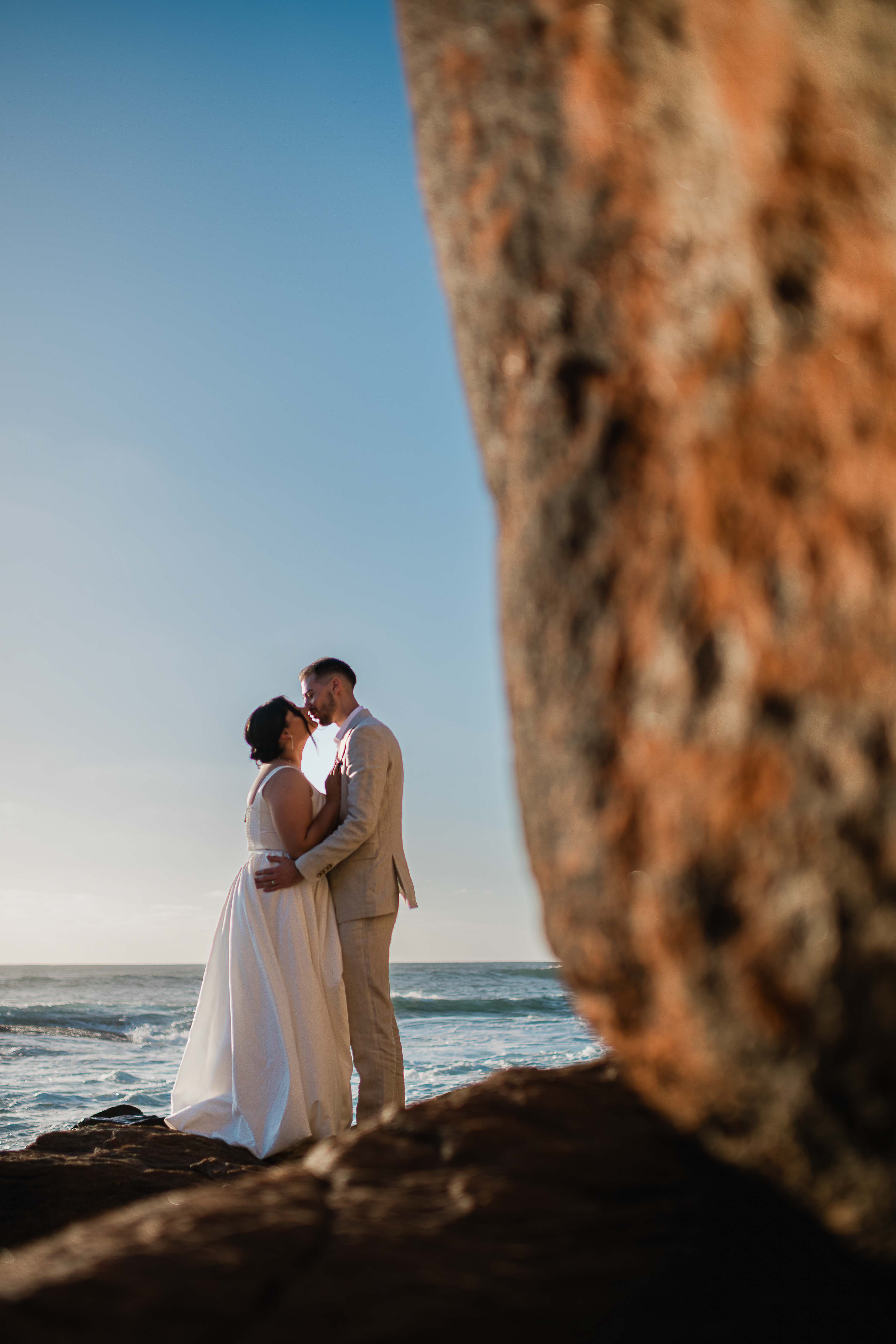 creative photo bride groom kissing framed by giant granite boulders leading the eye to the couple with golden hour ocean behind